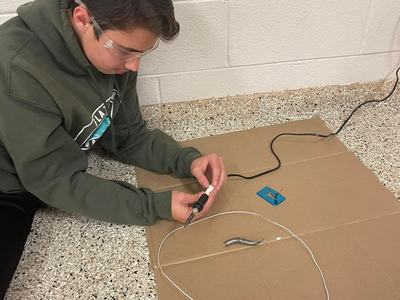 Person wearing safety glasses soldering a wire with a soldering iron on cardboard