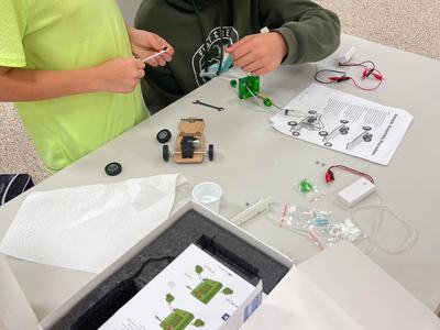 Two children assembling a small wheeled model car kit with instructions and parts on a table