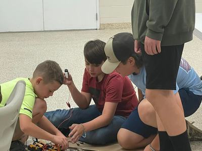 Three boys kneel on the floor assembling battery packs, wires, and components; another stands nearby.