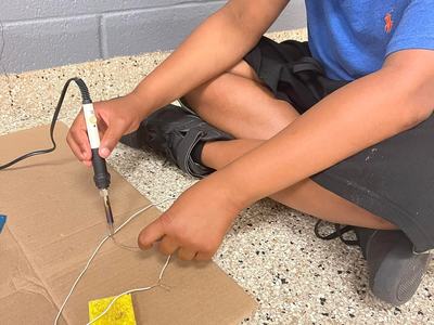 Student wearing safety goggles soldering two wires on cardboard while seated on floor
