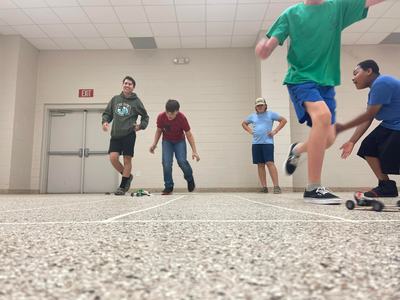 Boys racing small toy cars on a tiled floor inside a room, visible "EXIT" sign on wall