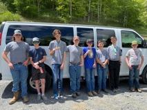 Group of people standing by white passenger van on gravel road with trees in background