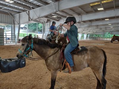 Child wearing helmet on horse in indoor arena holding a coiled lasso