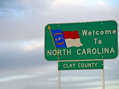 Green highway sign reading "Welcome To North Carolina" with state flag and "Clay County"
