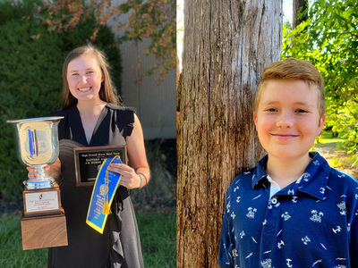 Young woman holding large trophy, plaque, and blue ribbon; boy standing beside a tree.