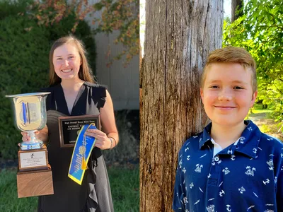 Young woman holding large trophy, plaque, and blue ribbon; boy standing beside a tree.