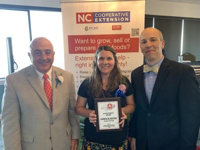 Three people posing at NC Cooperative Extension banner; woman holds plaque reading "Achievement Award Karen Blaedow State Winner"