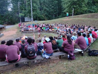 Large outdoor campfire gathering with campers seated on benches around fire, staff in maroon shirts