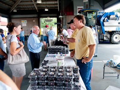 People buying packaged berries at a covered market stall with large farm machinery behind