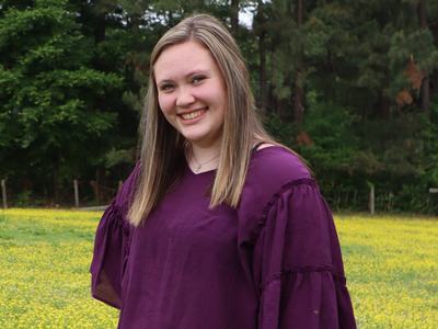 Young woman in purple blouse smiling while standing in a yellow wildflower field with trees
