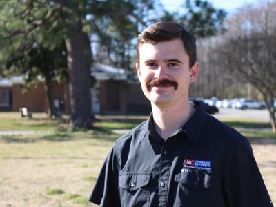 Man with mustache standing outdoors wearing shirt reading "NC Cooperative Extension"