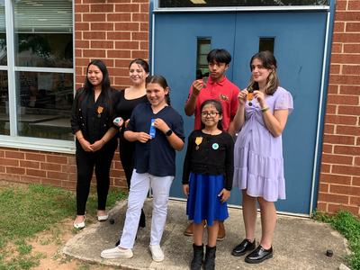 Six students standing outside a brick building by blue double doors holding ribbons and medals