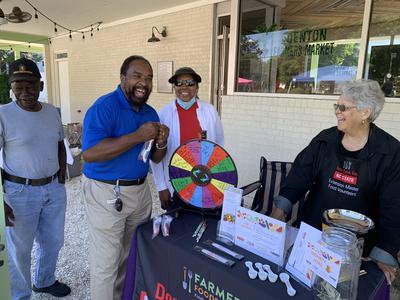 Four people at a farmers market booth with a colorful prize wheel and donation jar