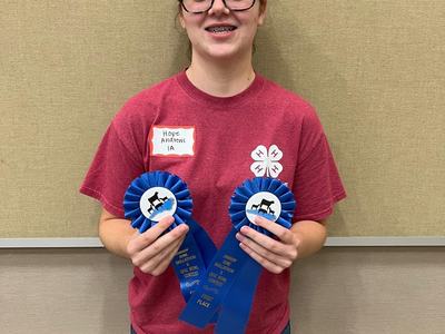 Person in red 4‑H shirt holding two blue ribbons labeled "First Place", name tag "Hope Andrews 1A"