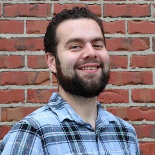 Bearded man in plaid shirt smiling in front of a red brick wall.