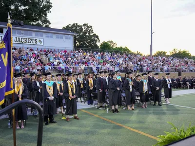 Graduating students in caps and gowns standing on a football field with audience in stands