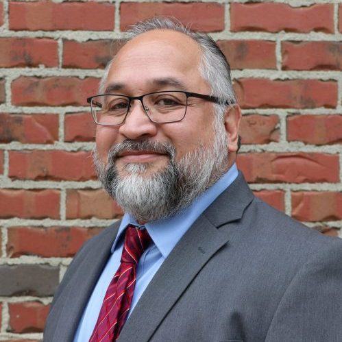 Bearded man with glasses in gray suit, blue shirt and red tie against a brick wall