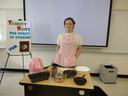 Person in pink apron behind table of cookware beside poster reading "Trinity Weitz: The Basics of Cooking! Yum"