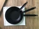 Three nested black frying pans with handles fanned on a white cloth on a wood table