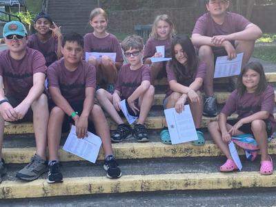 Children in matching maroon shirts sitting on outdoor steps holding papers