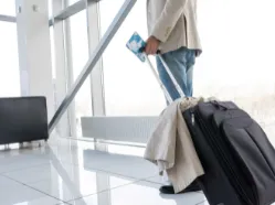 Traveler holding passport and boarding pass beside rolling suitcase with coat draped