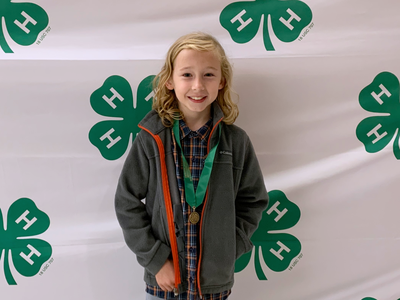 Young child wearing a medal standing in front of repeating 4‑H clover logos