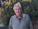 Older man standing in front of evergreen shrubs wearing a checked button-up shirt
