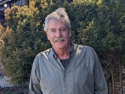 Older man standing in front of evergreen shrubs wearing a checked button-up shirt