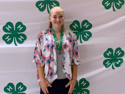 Person with medal standing in front of repeating green 4‑H clover logo backdrop