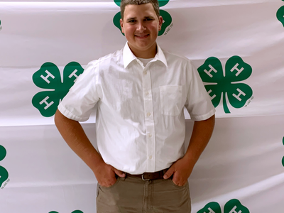 Teen wearing white shirt standing with hands in pockets in front of 4‑H clover backdrop