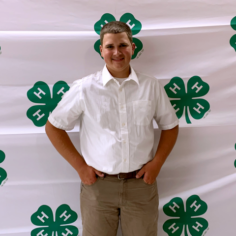 Teen wearing white shirt standing with hands in pockets in front of 4‑H clover backdrop