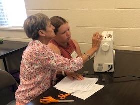 Older woman adjusts sewing machine while younger woman watches; papers and scissors on table.