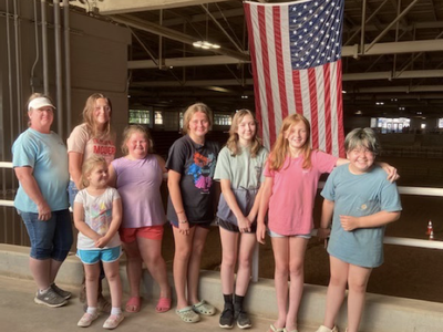 Group of eight girls and women standing indoors beside a large American flag