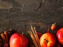 Row of red apples with pinecone, cinnamon sticks, wheat spikes and autumn leaves on dark slate