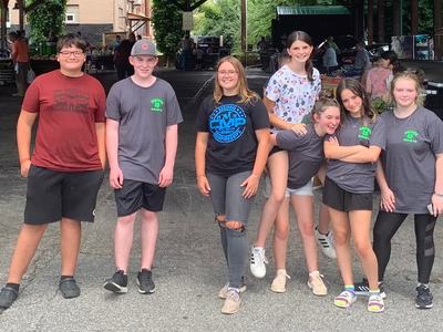 Seven teenagers standing under a pavilion at an outdoor market, posing for a photo