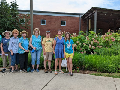 Group of nine people standing on a sidewalk in front of a brick building and garden