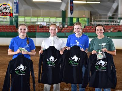 Four youth holding jackets labeled "NC State 4-H Horse Program" in indoor arena