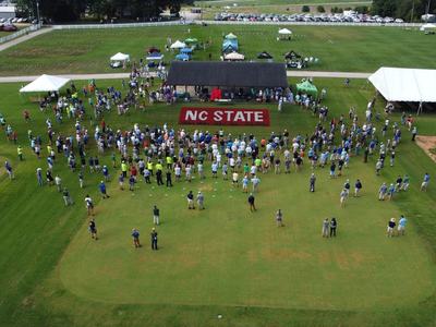 Drone aerial shot of Lake Wheeler Turf Field Lab, pavilion and turf painted "NC State" during 2022 Turfgrass Field Day. Attendees are clustered around the pavilion listening to announcements.