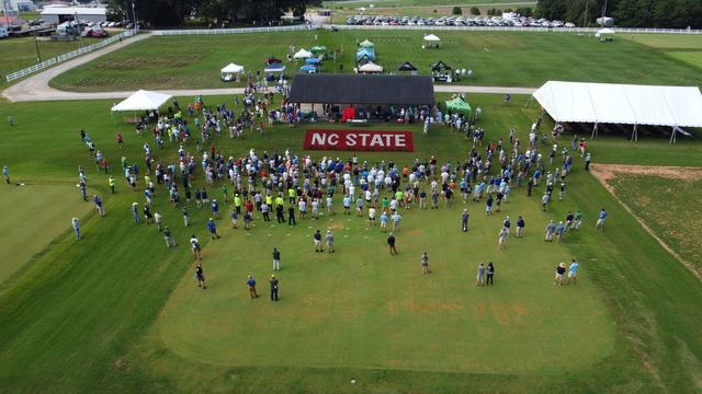 Drone aerial shot of Lake Wheeler Turf Field Lab, pavilion and turf painted "NC State" during 2022 Turfgrass Field Day. Attendees are clustered around the pavilion listening to announcements.