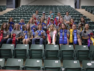 Group of young competitors seated in bleachers holding award ribbons (blue, red, yellow, pink)