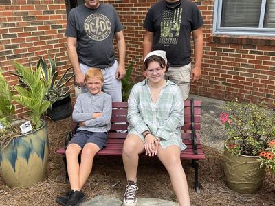 Four people posing on and behind a bench outside a brick building