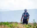 Man in cap and gloves with safety harness walking in grassy field holding a tool