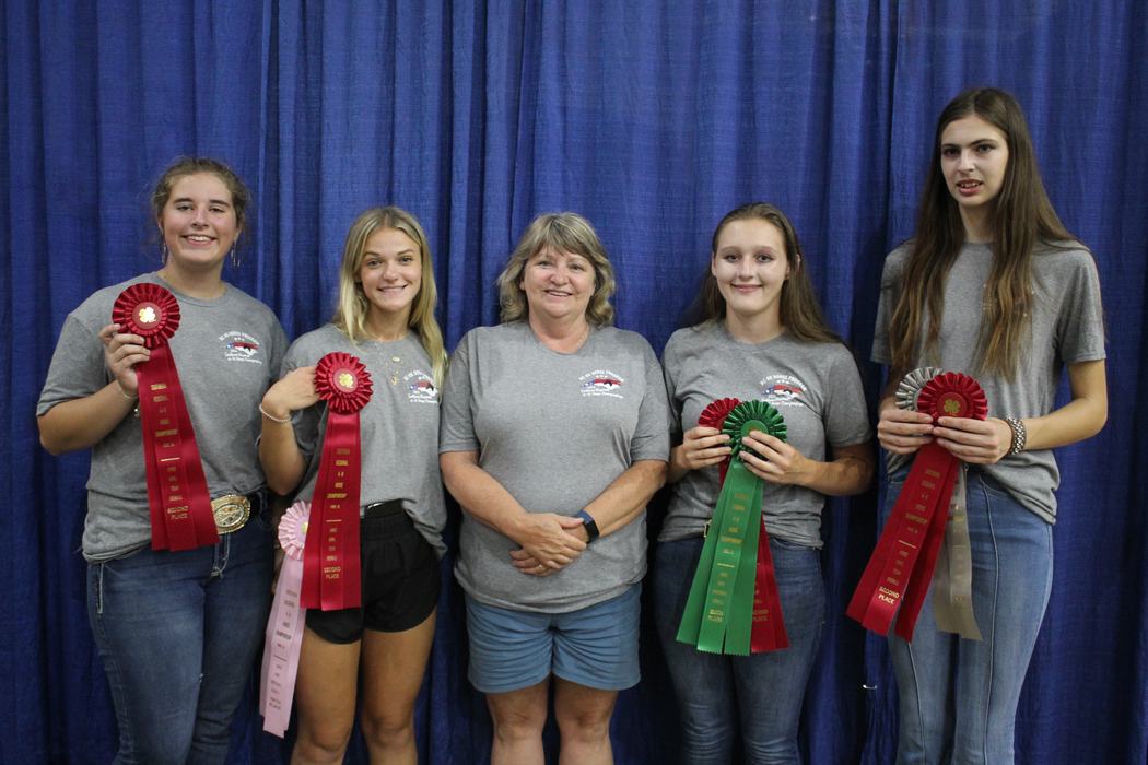 Five people standing before a blue curtain holding red, green, pink, and beige award ribbons
