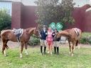 Two equestrians in riding gear with horses and a woman standing in front of 4‑H clover sign