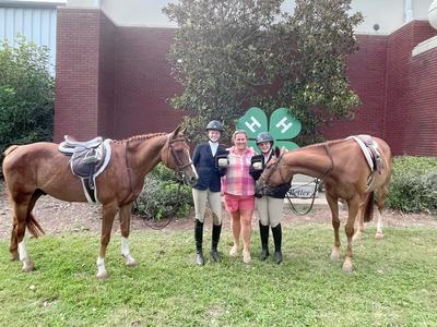 Two equestrians in riding gear with horses and a woman standing in front of 4‑H clover sign