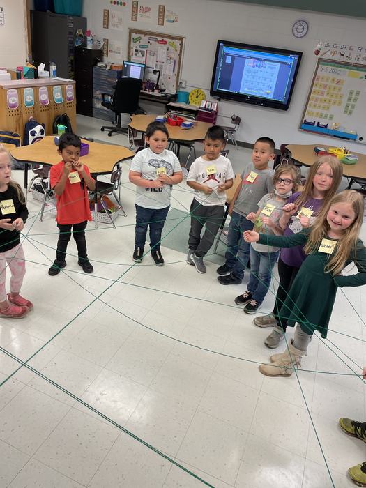 Elementary students standing in a circle holding a green string web; whiteboard reads "4-5-2022".