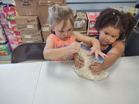 Two children squeeze and seal a zip-top plastic bag containing dough or batter at a table.