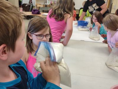 Children at table mixing dough in zip-top bags, teacher nearby wearing "ALL DAY EVERY DAY" shirt