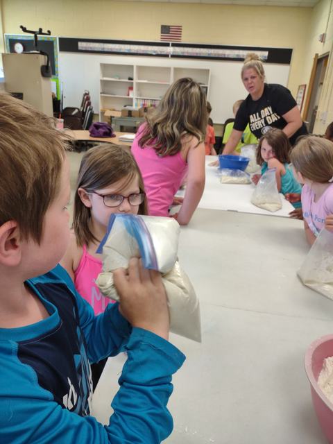 Children at table mixing dough in zip-top bags, teacher nearby wearing "ALL DAY EVERY DAY" shirt