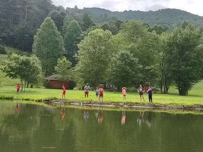 Group of people standing along a pond shoreline in grassy, tree-lined mountain area.